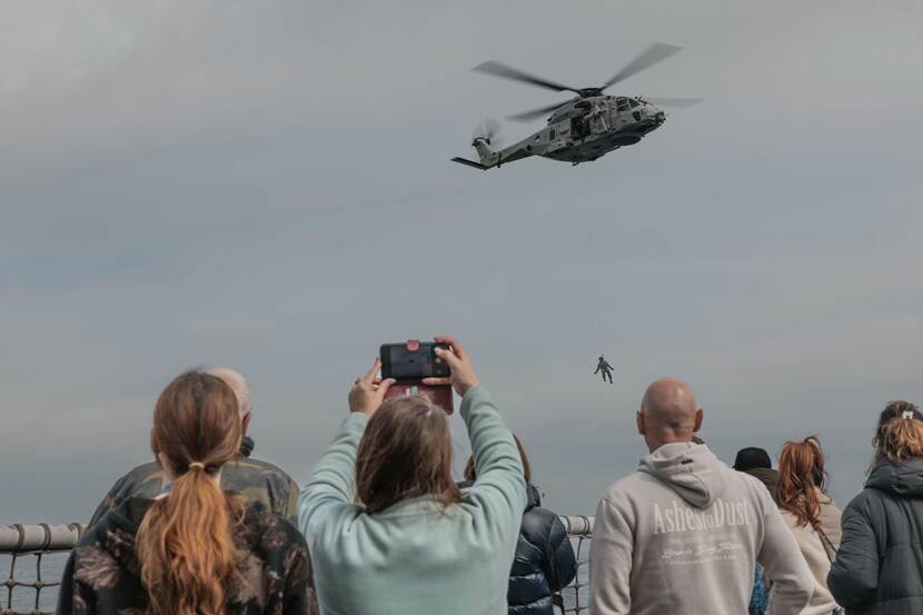 Vanaf schip kijken mensen naar NH90 helikopter met militair die aan touw uit toestel hangt.