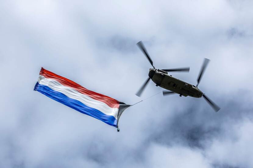 Chinook-transporthelikopter vliegt met Nederlandse vlag eronder.