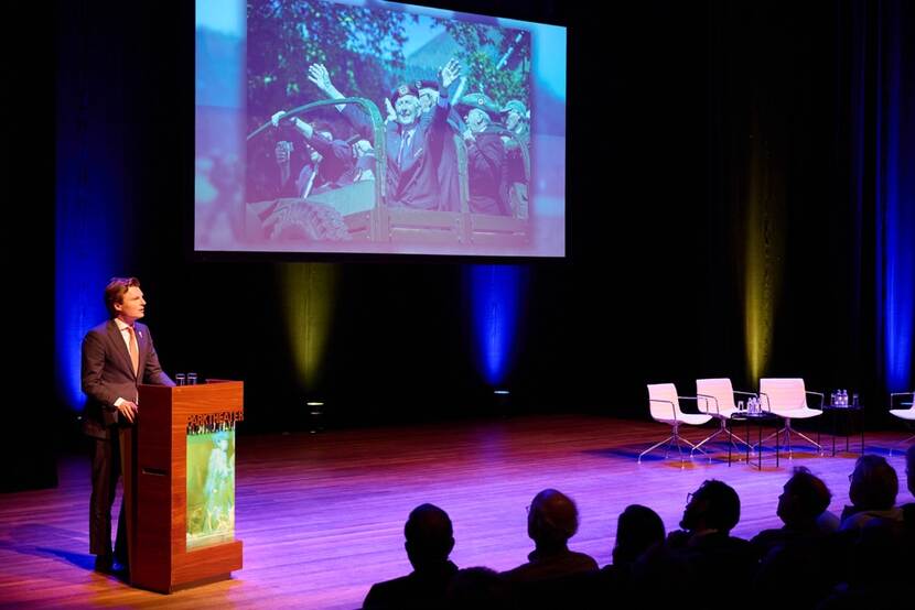 Minister Brekelmans staat achter het spreekgestoelte. Op een scherm achter hem is een foto van veteranen te zien.