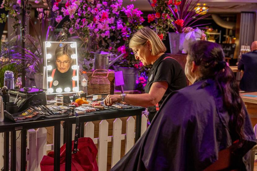 Een mevrouw wordt opgemaakt. In spiegel reflecteert gezicht visagiste. Paarse bloemen op achtergrond.