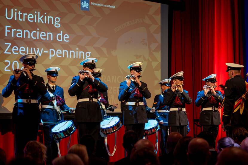 Militair muziekkorps speelt. Op de achtergrond scherm met 'Uitreiking Francien de Zeeuw-penning'