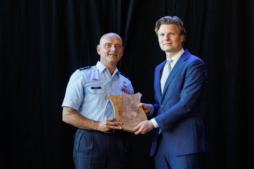 Generaal Steur (in luchtmachtpak) houdt een houten embleem in handen, samen met minister Brekelmans op rechts, die een pak draagt.