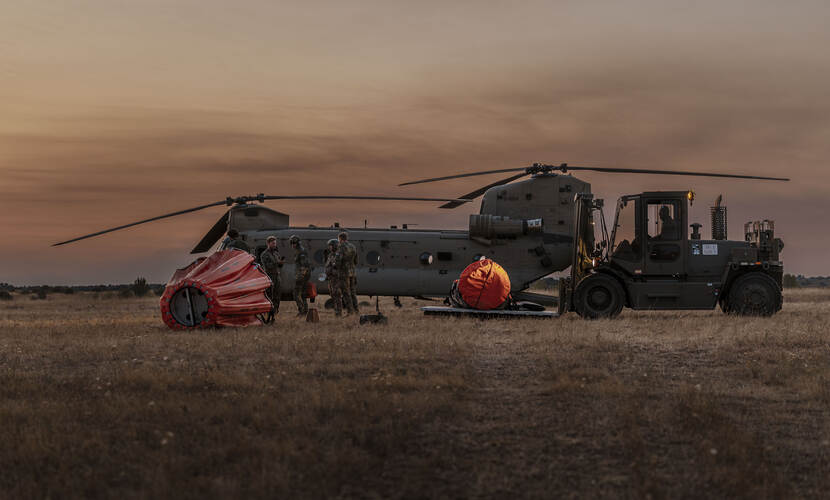 Chinook-helikopter staat weer aan de grond. Oranje bambi bucket ligt ernaast, met 4 militairen erbij. Rechts een heftruck.