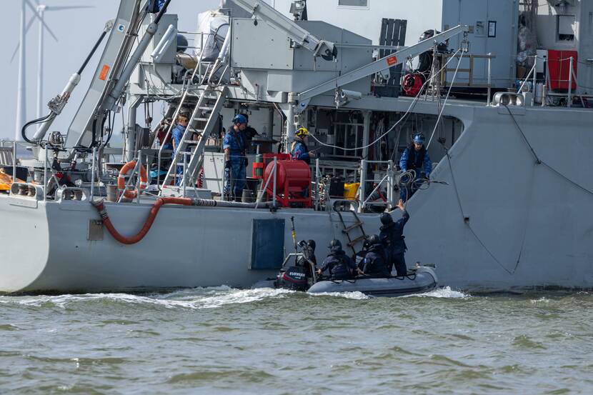 4 zwartgeklede personen in een rubberboot bij een trapje dat naar een groter schip leidt.