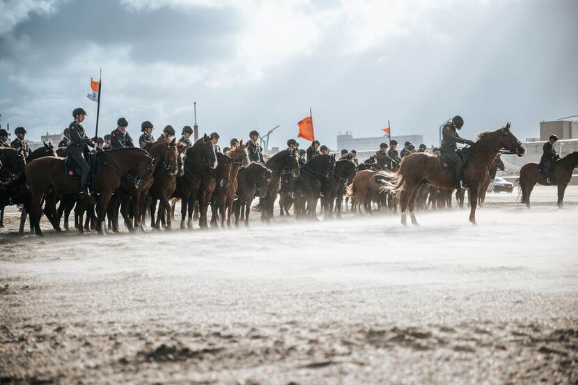 Een 30-tal paarden met berijders erop trotseren op het strand een windvlaag met strandzand.