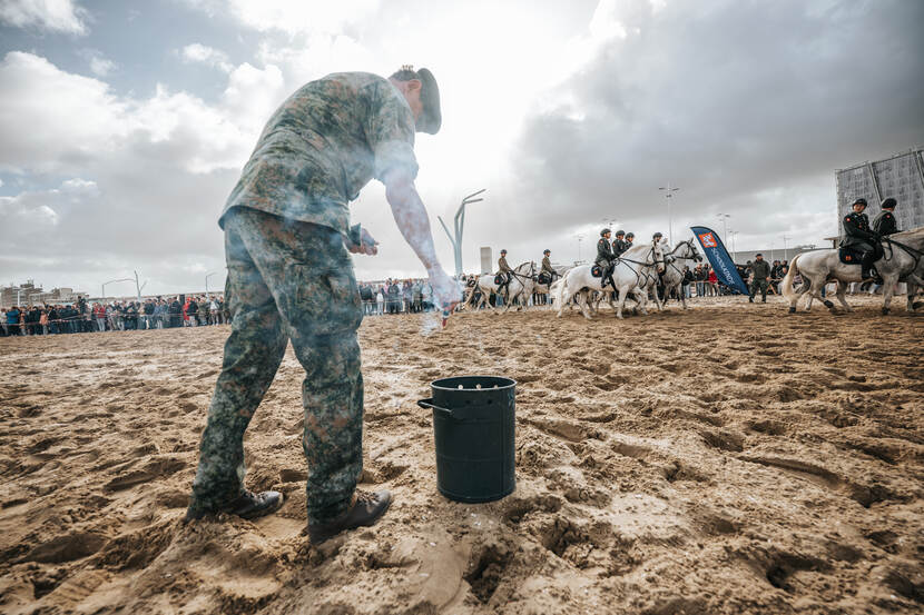 Tijdens de generale repetitie voor Prinsjesdag steekt een militair op het strand knal- en rookpotten af. Zijn rechterarm is bedekt met rook. Op de achtergrond toeschouwers en ruiters met paarden.