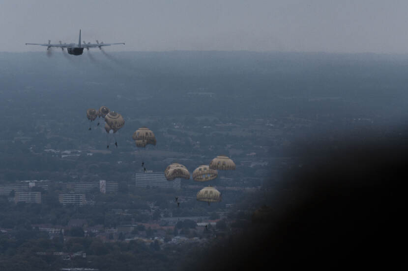 Parachutisten hangen aan hun parachutes onderweg naar beneden. Linksboven op de foto het vliegtuig waar ze net uit zijn gesprongen.