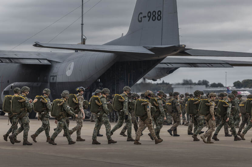 Hercules met op de voorgrond een twintigtal militairen die met een parachute op hun rug lopen.