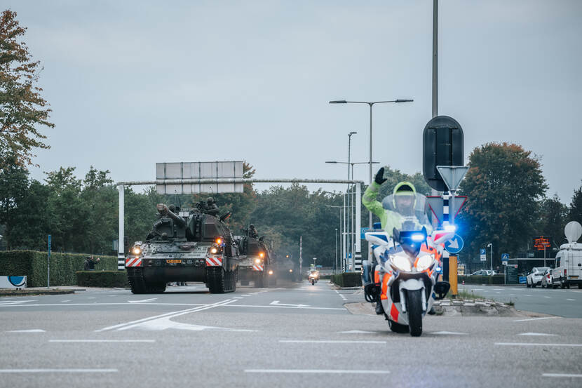 Een militaire colonne rijdt over de weg met voorop een motor van de Koninklijke Marechaussee.