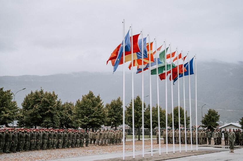 Vlaggen van 11 landen wapperen op lange masten. Daarvoor staan rijen dik militairen.