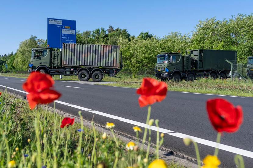 3 militaire vrachtwagens draaien een asfaltweg op. Rode klaprozen op de voorgrond.