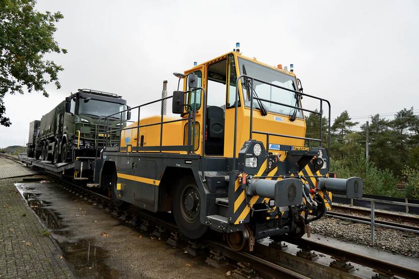 Groene Defensievrachtwagens op een oplegger op het spoor met een gele locomotief ervoor.
