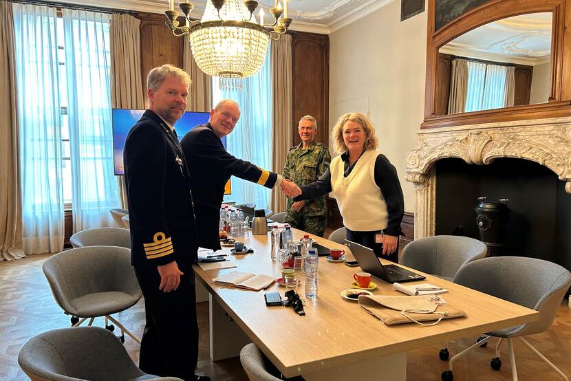 3 militairen in uniform en 1 vrouw staan rond een tafel. De vrouw in burgerkleding en een militair schudden de hand.