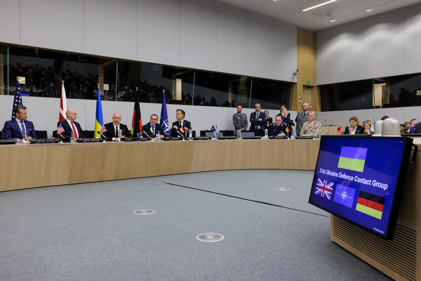 Zaal met sprekers van de 31e Ukrain Defence Contact Group. Op de voorgrond is veel grijze vloer zichtbaar.