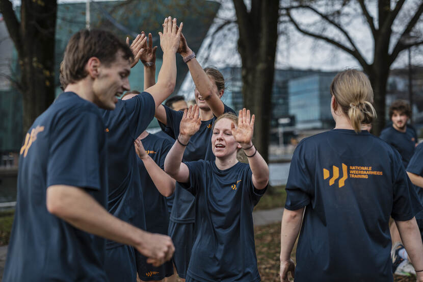 Een 8-tal jongeren met een T-shirt aan met de tekst Nationale Weerbaarheids Training erop. Met de armen omhoog klappen  sommigen tegen de handen van een ander.