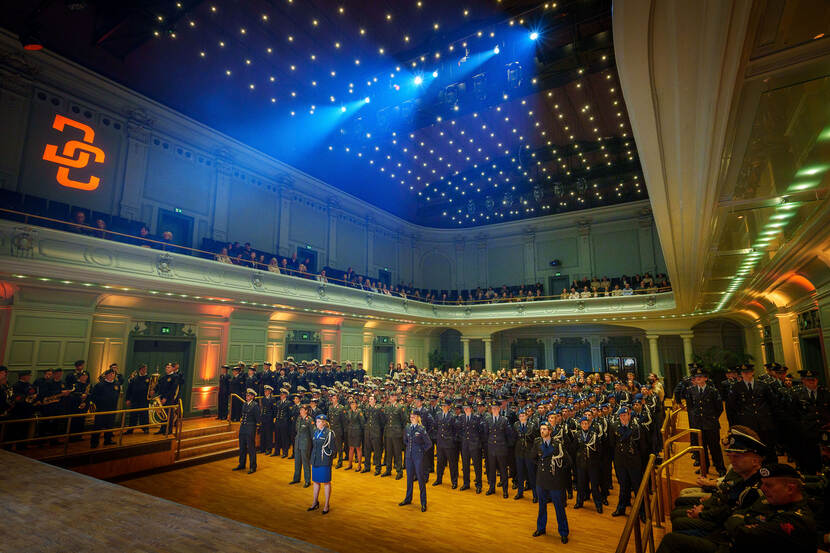 Een zaal met enkele honderden militairen in uniform die richting podium kijken. Het logo van Defensity College, DC, wordt op de muur geprojecteerd.