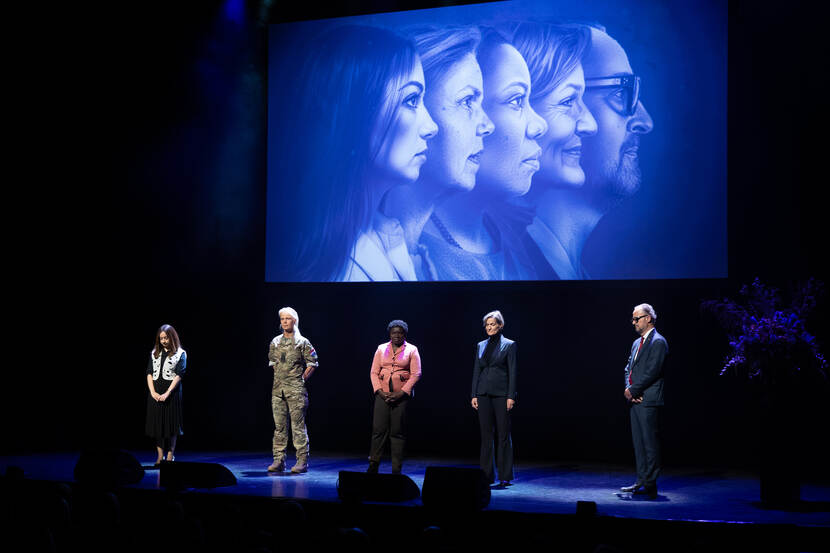 Deelnemers aan de podcast staan op een podium. Achter hun zijn hun gezichten te zien op een scherm.