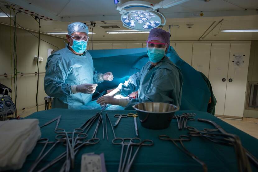 2 mensen van een medisch team in groene kleding met mondkapje en hoofdbedekking op bezig met een patient op een marineschip.