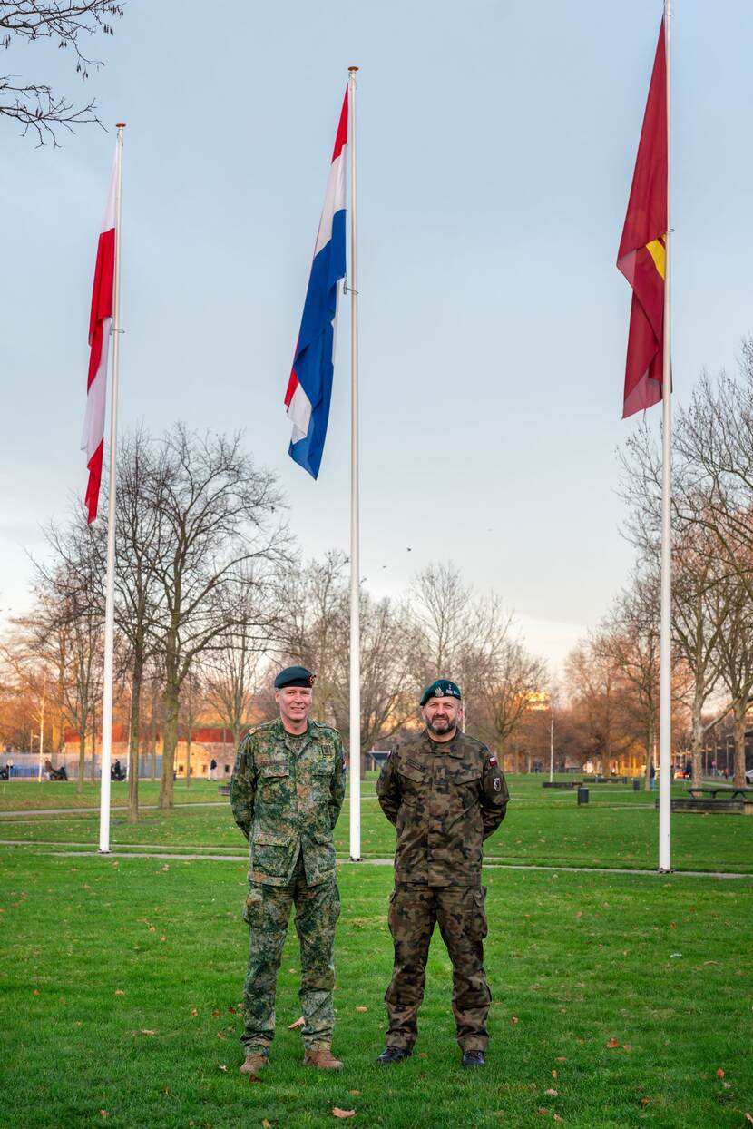 2 militairen op een grasveld met 3 vlaggen op de achtergrond.
