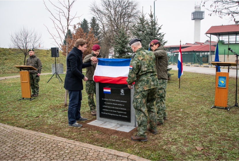 De minister en de CDS halen de Nederlandse vlag omhoog waarna een gedenksteen zichtbaar is geworden. Ze staan op een grasveld. De lucht is grijs.