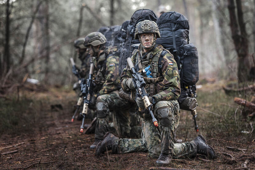 Deelnemers aan de opleiding zitten met bepakking op op hun knieën in het bos.