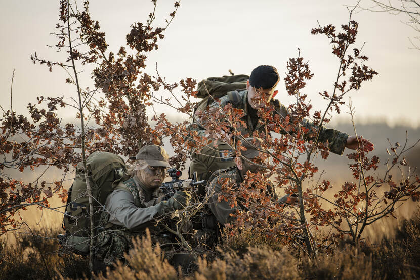 Amalia zit gecamoufleerd tussen de struiken met een geweer.