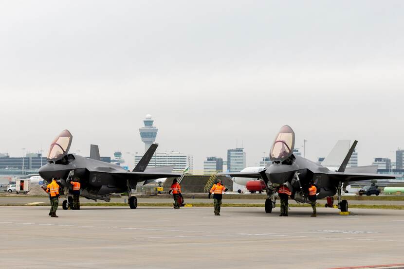 2 F-35's staan geparkeerd op Luchthaven Schiphol. Eromheen staan en lopen 7 mensen met reflecterende hesjes.