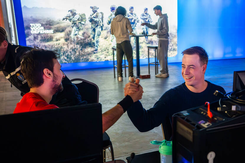 2 jonge mannen geven elkaar een high five. Ze zitten achter een beeldscherm. Op de achtergrond 2 anderen die aan een statafel een potje schaken.