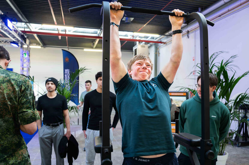 Een jonge man trekt zich op aan zijn armen aan een staaf. Hij heeft een vertrokken gezicht.