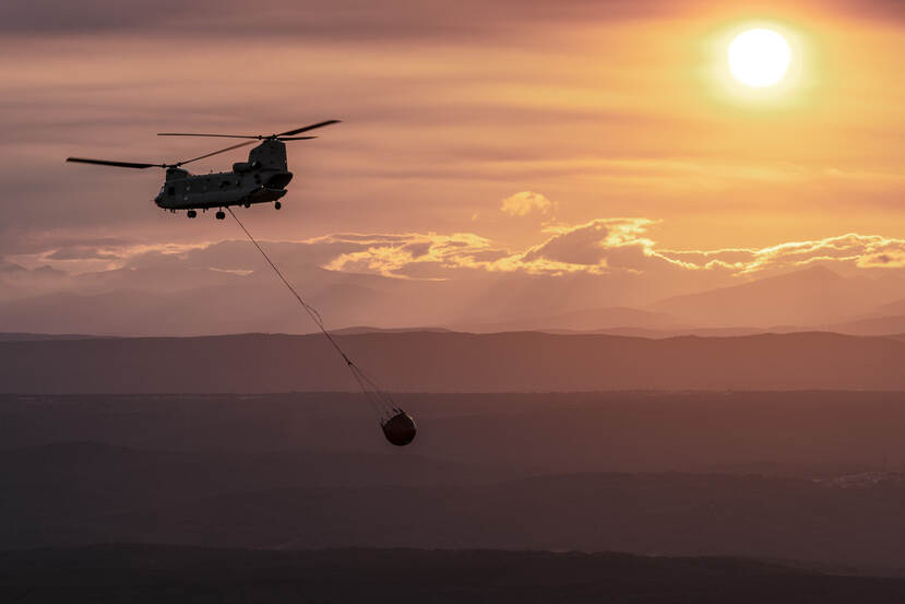 Een Chinook in de lucht met bambi bucket aan een touw eronder met een zalmroze achtergrond van zon, een laag wolken eronder en heuvelachtig terrein.