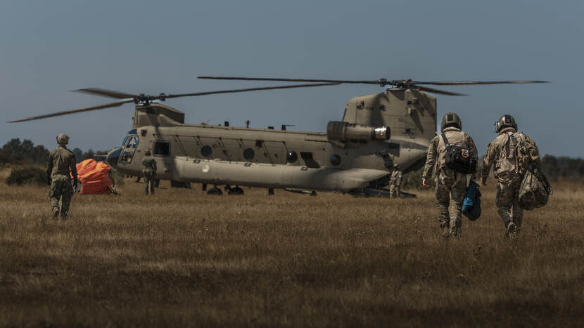 4 militairen op de rug gezien die naar een Chinook-transporthelikopter lopen. Links bij de neus van de helikopter de oranje bambi bucket waarin water wordt vervoerd.