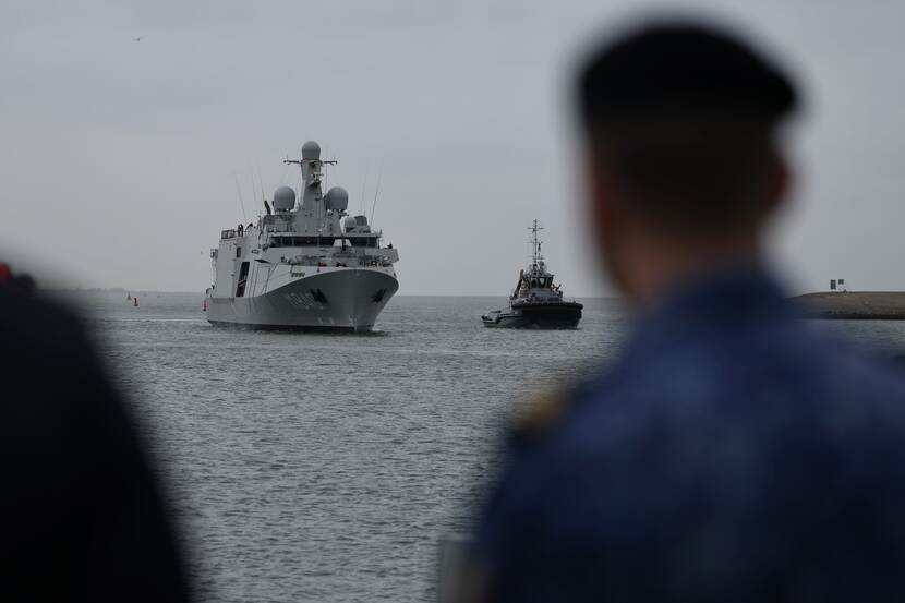 Het grijze schip De Vlissingen komt aangevaren, rechts geflankeerd door een sleepbootje. Rachts vaag de contouren van een marinier.