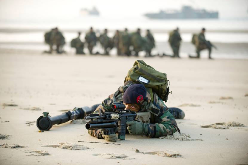Marinier kijkt door vizier, liggend op het strand.