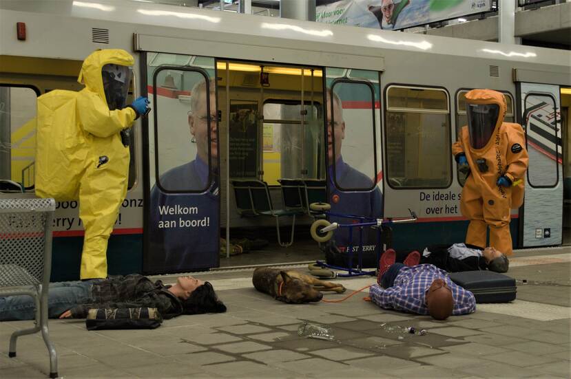 Gewonde mensen liggen op metroperron. CBRN-specialist in geel pak neemt een monster.