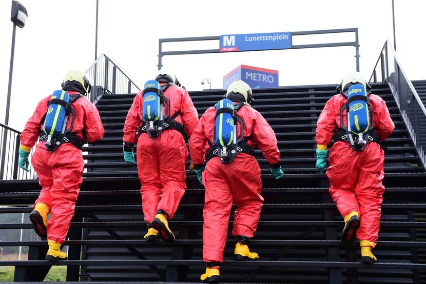 Personen in rode beschermende pakken en gasflessen op de rug lopen trap van een metrostation op.