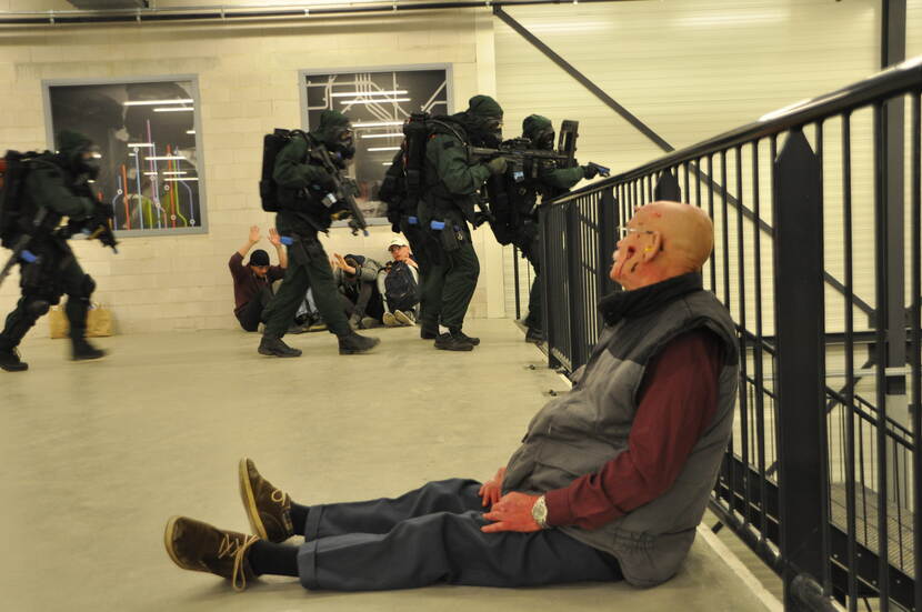 Gewonde man zit tegen een hek in een metrostation. Personen in beschermende pakken lopen een trap af.