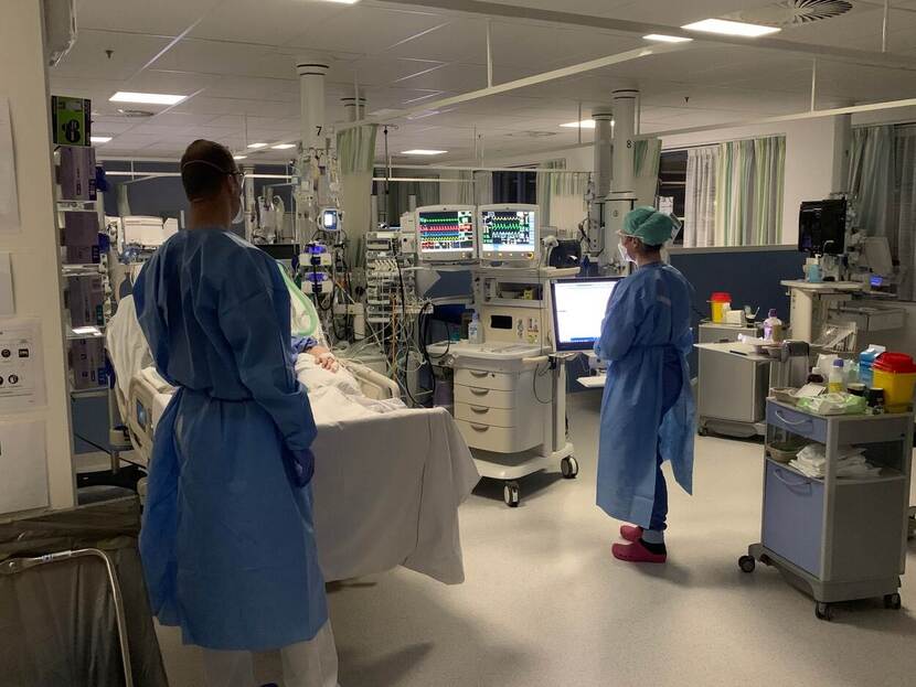 2 personen met blauwe ziekenhuiskleding staan naast het bed van een patiënt.