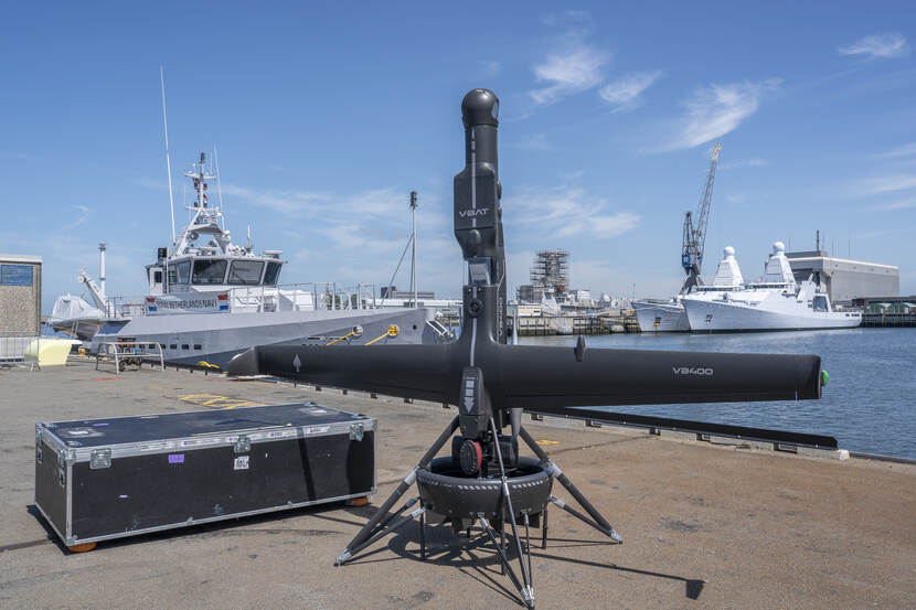 De zwarte V-BAT staat rechtop. Daarachter liggen marineschepen aan de kade.