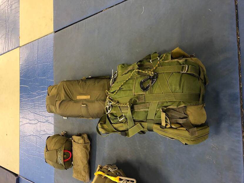 Ingepakte parachute met militaire bepakking aan aflaatsysteem op blauwe sportmatten.