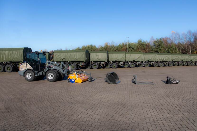 Wiellaadschop op een parkeerplaats met aanhangwagens met verschillende opzetstukken waaronder een borstel, palletvork en puinbak.