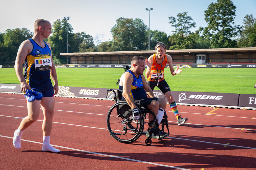 Een man in een roelstoel op de atletiekbaan wordt aangemoedigd door een dame.