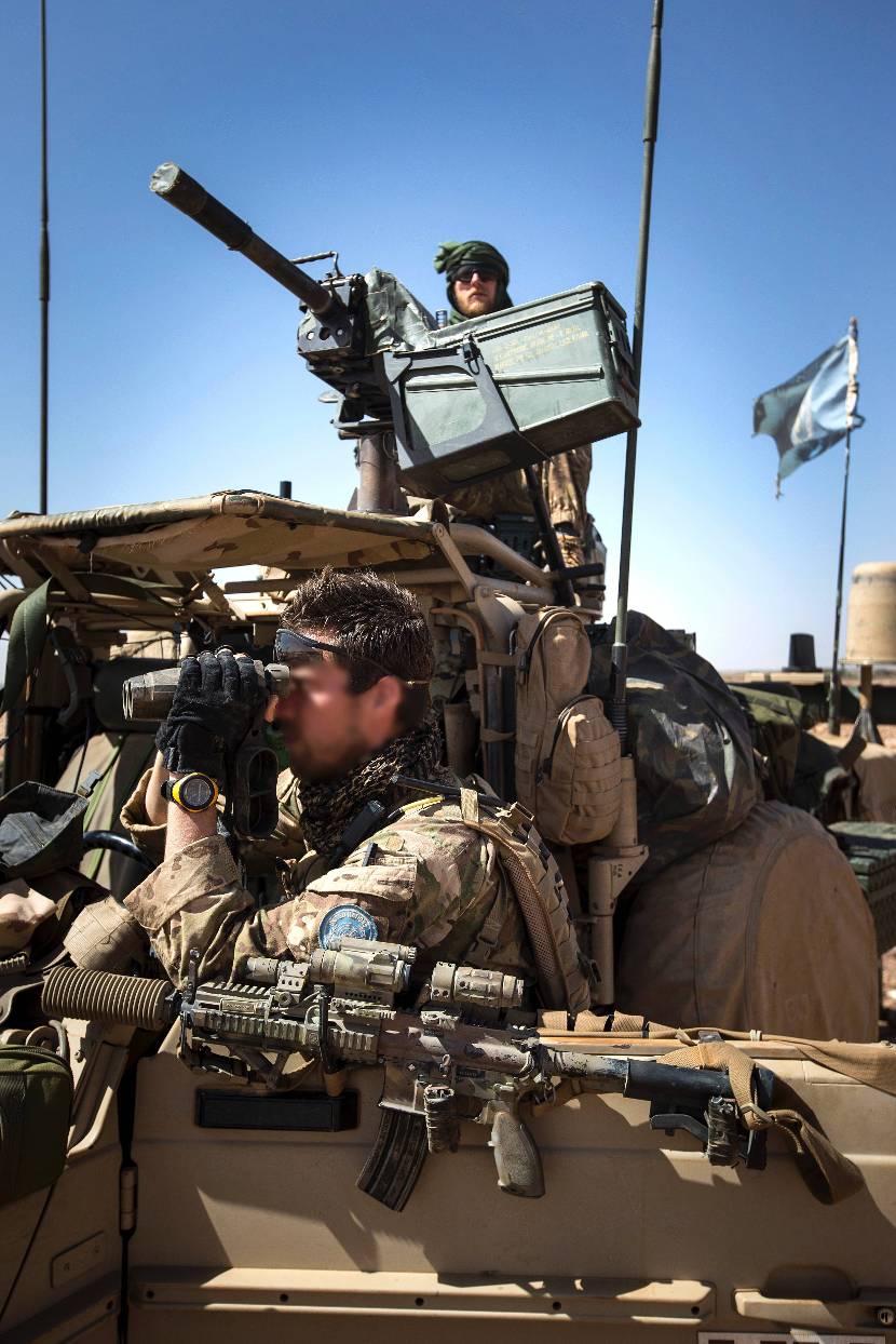 Militair op een voertuig houdt vanachter een automatische granaatwerper de omgeving in de gaten. Op de voorgrond een militair die door een verrekijker kijkt.