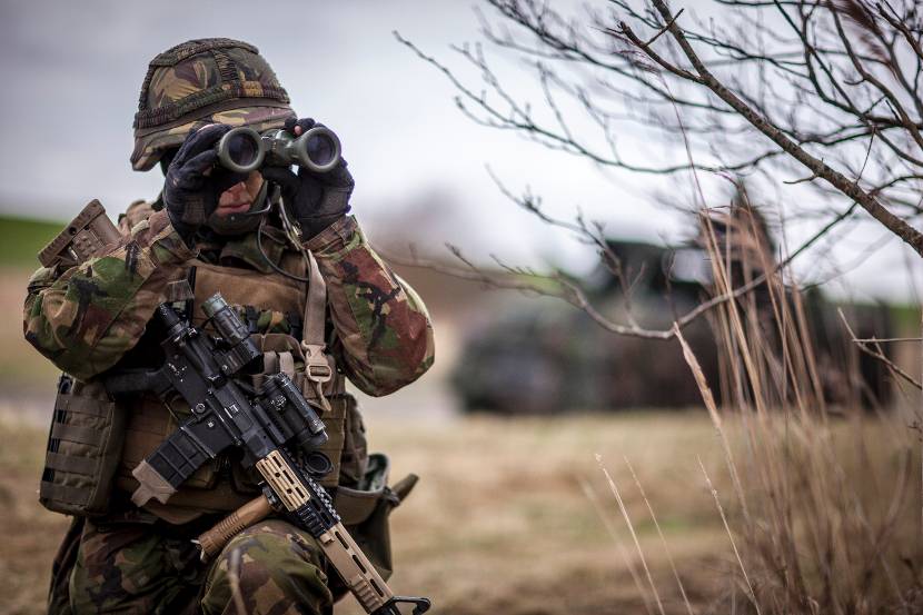 Militair kijkt door een verrekijker. Een Colt C7-geweer steunt op z'n linkerknie.