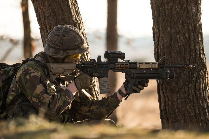 Een militair met een Colt C7-geweer zittend tussen bomen.