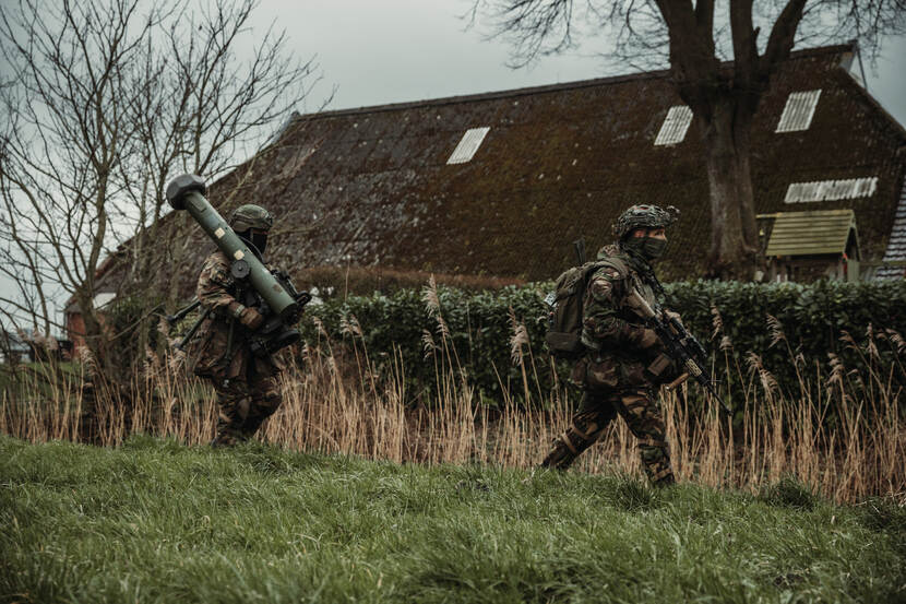 2 militairen lopen met wapens waaronder een Spike LR-antitankwapen door het gras langs een boerderij.