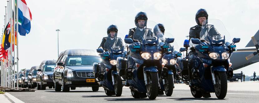 Motoren marechaussee begeleiden rouwwagens vanaf platform met militaire transportvliegtuigen. Vlaggen verschillende landen halfstok.