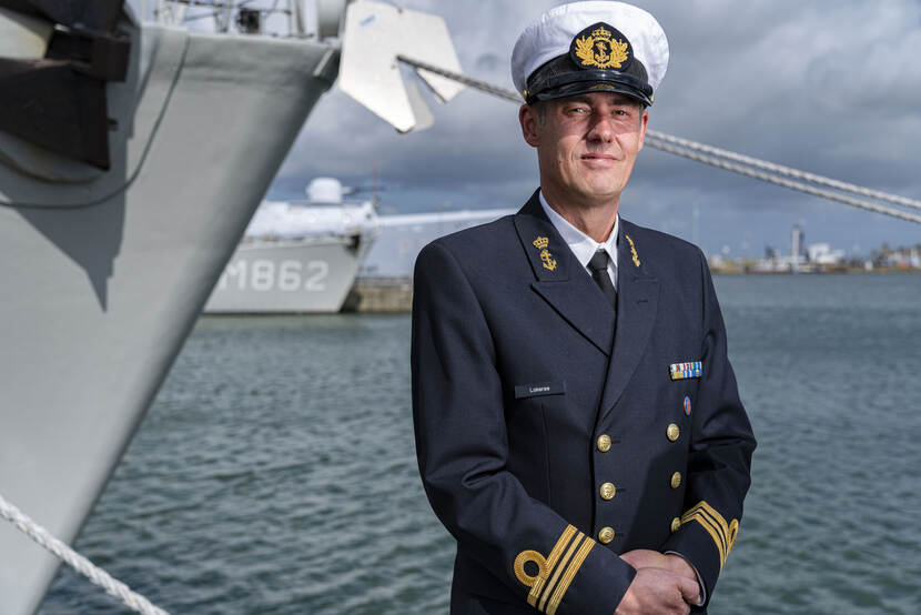 Marineofficier geportretteerd voor 2 marineschepen in de haven.