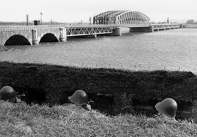Zwart-wit foto van 3 militairen tijdens Meidagen 1940. Ze liggen in dekking bij brug over water.