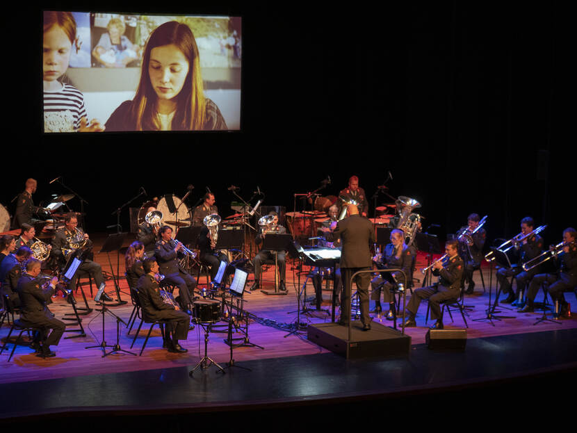 Een militaire orkest speelt muziek met op de achtergrond een beeldscherm. Daarop een mevrouw en een kind.