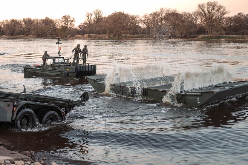 militairen staan op een boot. Tegenover hen ontvouwt drijvend platform. Aan de kant van het water staat een voertuig deels in het water.